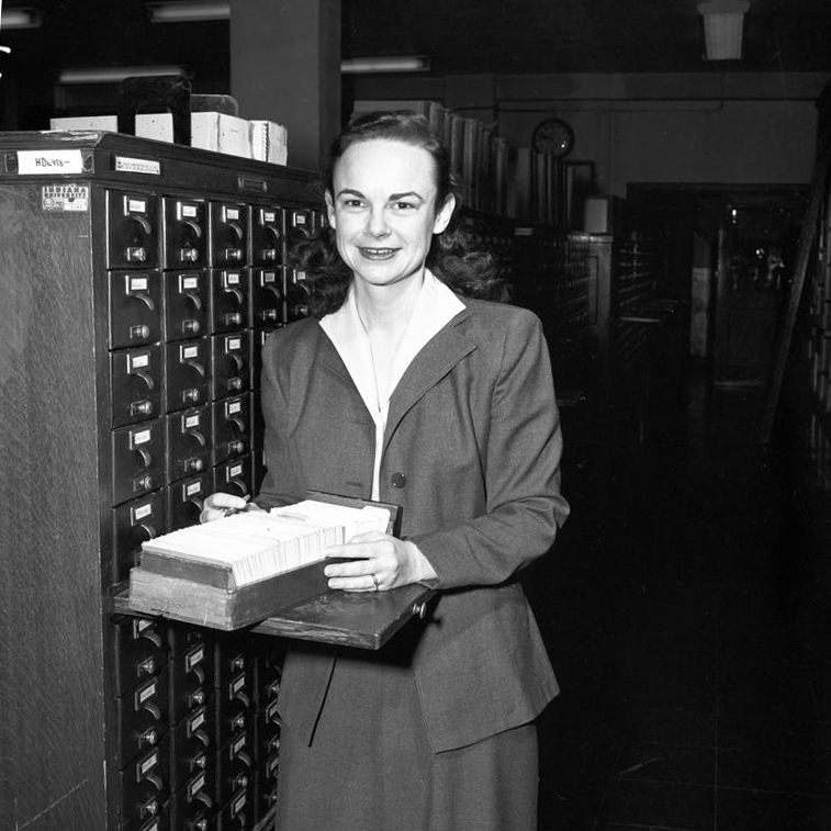 Library staff member Simone Robbins stands beside a card catalog smiling at the camera. A card file rests before her. Photo taken on February 10, 1954.