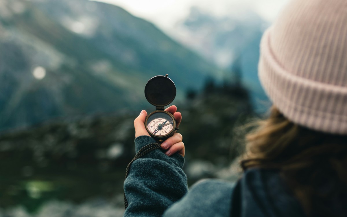 In the foreground, a person holds a compass in their hand. In the background, tree-covered mountains are visible.